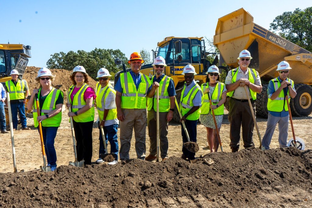 Caring Hands leadership, board members, and community partners in hard hats and safety vests turning the first shovels of dirt at the groundbreaking ceremony, August 14, 2026.